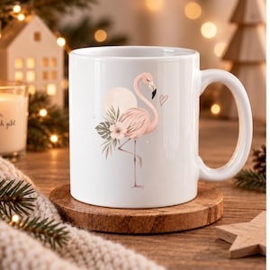 Puede incluir: Una taza de cerámica blanca con una ilustración impresa de un flamenco rosa claro de pie sobre una pata. El diseño incluye un sol circular pálido, hojas tropicales verdes, una pequeña flor y el contorno fino de un corazón junto al flamenco.