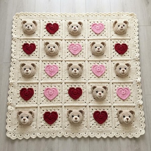Könnte beinhalten: Eine quadratische Häkeldecke aus cremefarbenem Garn mit einem Gitter aus abwechselnden Quadraten. Die Quadrate zeigen entweder ein dreidimensionales hellbraunes Teddybärengesicht oder ein flaches Herz in Rot- und Rosatönen mit Bogenkante.