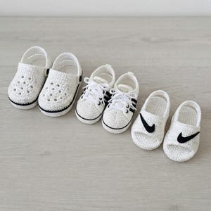 May include: Three pairs of white crocheted baby shoes. The first pair are clogs with perforated tops and black trim. The second pair are lace-up sneakers with black side stripes. The third pair are open-toe sandals with black swoosh logos.