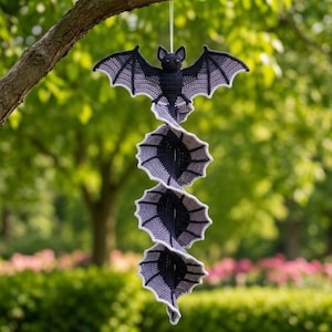Puede incluir: Un móvil de viento tejido a ganchillo que presenta un murciélago de color gris oscuro y gris claro en la parte superior con las alas extendidas, seguido de una serie de tres segmentos en espiral con forma de alas de murciélago a juego.