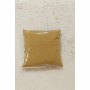 Puede incluir: Una bolsa cuadrada de plástico transparente llena de un polvo fino de color amarillo mostaza. El polvo dentro de la bolsa sellada muestra una red de finas fisuras oscuras en su superficie, creando una apariencia texturizada en el empaque.