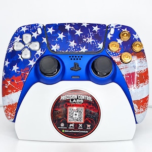 May include: A wireless game controller featuring a distressed American flag pattern in red, white, and blue. It has silver directional buttons, gold action buttons, and black joysticks. The controller rests on a white stand with a circular logo.