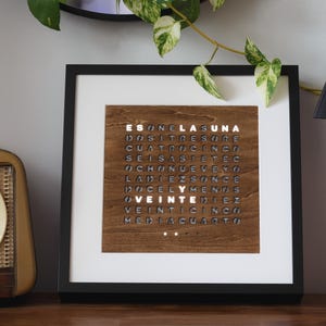 Könnte beinhalten: Eine quadratische Wortuhr in einem schwarzen Rahmen mit einer dunklen Holzfront, die ein Gitter aus spanischen Buchstaben zeigt. Die Wörter ES, LA, UNA und VEINTE leuchten hellweiß vor dem dunklen Holzhintergrund und zeigen die Uhrzeit an.