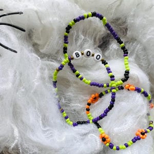 Op de afbeelding: Twee kralenarmbanden met kleine ronde kralen in paars, limoengroen, zwart en oranje. Eén armband bevat witte letterkralen die het woord BOO spellen, terwijl de andere armband is voorzien van kleine oranje bloemvormige kralen tussen de rest.