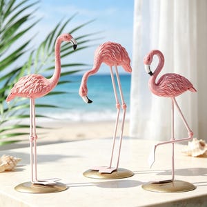 May include: Three pink flamingo figurines standing on circular gold bases. Each flamingo features textured feathers, long thin legs, and a beak with a black tip. The figures are posed in different positions, with one standing tall, one bending down, and one standing on one leg.
