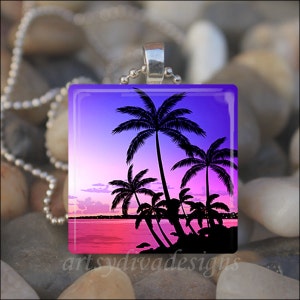 May include: A glass pendant necklace with a silver chain features a colourful sunset scene with palm trees silhouetted against the sky. The sky is a gradient of purple, pink and orange.