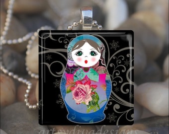 Puede incluir: Un colgante de vidrio con una muñeca rusa estilizada de colores con un diseño floral y remolinos negros y plateados en el fondo.