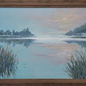 May include: An oil painting in a brown wooden frame depicting a calm lake at dawn or dusk. The scene features soft blue and pink hues in the sky and water, with dark silhouettes of trees on the distant shore and tall grasses in the foreground.