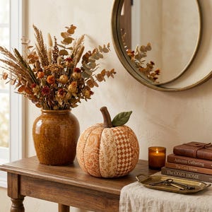May include: A brown ceramic vase filled with dried grasses and flowers, a fabric pumpkin with mixed orange and cream patterns, a small glass candle, and a stack of three books titled Autumn Gathering and The Cozy Home on a wooden table.