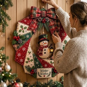 May include: A quilted Christmas wreath featuring red, green, and cream fabric panels with snowflake and holly patterns. A fabric snowman wearing a top hat and scarf hangs in the center, holding a lantern. A sign at the bottom reads Winter Wishes.