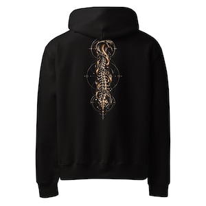 Può includere: Una felpa con cappuccio nera con una grafica color oro sul retro. Il design raffigura una colonna vertebrale verticale intrecciata con un cobra, circondata da cerchi geometrici e dal testo THE STRENGTH REMAINS in formato striscione.