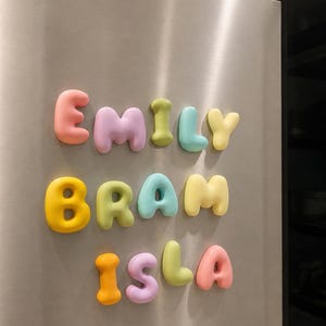 Puede incluir: Un conjunto de letras magnéticas tridimensionales de colores pastel dispuestas en tres filas sobre una superficie de acero inoxidable. Las letras forman los nombres EMILY, BRAM e ISLA con caracteres redondeados tipo burbuja en tonos rosa, amarillo, verde y azul.