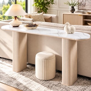 May include: A long, narrow console table with a white faux marble top and two light-colored, vertically fluted cylindrical bases. A matching fluted, round ottoman sits underneath the table between the two bases.