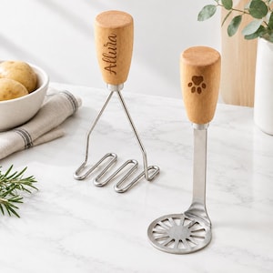 May include: Two kitchen mashers with light wood handles. One masher features a wire frame and the name Allura engraved on the handle. The other masher has a flat, circular metal base with radial slots and a paw print engraved on the handle.