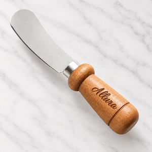 May include: A butter spreader with a silver-toned metal blade and a cylindrical wooden handle. The handle features a rounded bolster and is engraved with the word Allura in a cursive script. The spreader has a smooth, polished finish.