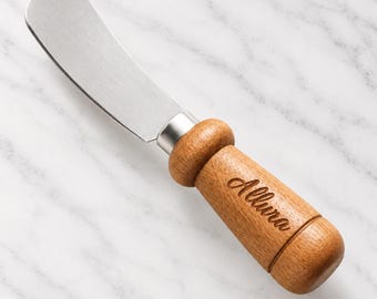 May include: A butter spreader with a silver-toned metal blade and a cylindrical wooden handle. The handle features a rounded bolster and is engraved with the word Allura in a cursive script. The spreader has a smooth, polished finish.