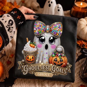 May include: A black t-shirt featuring a graphic of a rhinestone-covered ghost with a large colorful bow, holding a pumpkin-themed drink and a jack-o'-lantern bucket. Below the ghost is a gold banner with the text GHOUL-IEST GOALS and GHOUL.