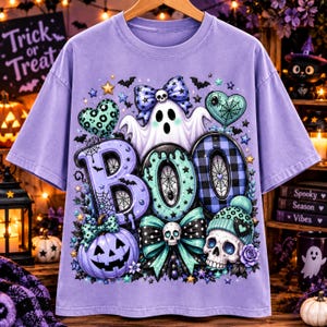 Puede incluir: Una camiseta de color morado claro con un estampado gráfico que muestra la palabra BOO en letras grandes. El diseño incluye un fantasma blanco, una calabaza morada, una calavera con gorro, lazos, corazones, estrellas y murciélagos en tonos morados.