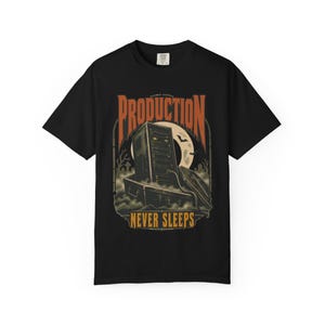 May include: A black short-sleeve t-shirt featuring a graphic print of a computer tower rising from a coffin. The text reads PRODUCTION NEVER SLEEPS in orange and yellow lettering. The background includes a full moon, bats, and a graveyard.