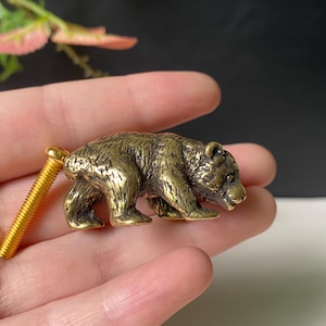 Könnte beinhalten: Ein messingfarbener Möbelknopf in Form eines gehenden Bären mit detaillierter Fellstruktur. Die Bärenfigur ist an einer langen, goldfarbenen Metallschraube mit Gewinde befestigt, die an der Rückseite herausragt und zur Montage dient.