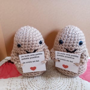 Könnte beinhalten: Zwei beige gehäkelte Kartoffelfiguren mit schwarzen Kunststoffaugen und gestickten Lächeln. Jede Figur hält eine weiße Karte mit dem französischen Text 'Je suis une petite patate positive' und 'Je crois en toi' über einem kleinen roten Herz.