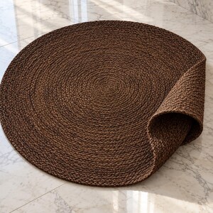 May include: A circular rug crafted from dark brown braided fibers, featuring a coiled pattern that radiates from the center. One edge of the rug is folded upward, revealing the underside and the thickness of the woven material construction.