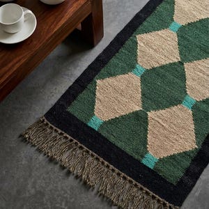 May include: A rectangular woven runner rug featuring a geometric diamond pattern in shades of forest green, beige, and teal. The rug is framed by a solid black border and finished with long, textured brown fringe tassels on the short ends.