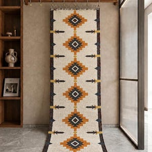 May include: A long, rectangular woven wall hanging featuring a cream-colored background with a repeating geometric pattern in mustard yellow, charcoal gray, and black. The textile has dark brown side borders and fringed edges at the top and bottom.