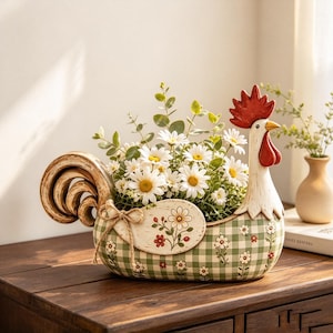 Könnte beinhalten: Ein Keramik-Pflanzgefäß in Hahnform mit grün-weißem Vichy-Muster, verziert mit kleinen gemalten Blumen und einer Juteschleife. Das Gefäß ist mit weißen Gänseblümchen und grünen Eukalyptusblättern gefüllt und hat einen roten Kamm sowie rote Kehllappen.