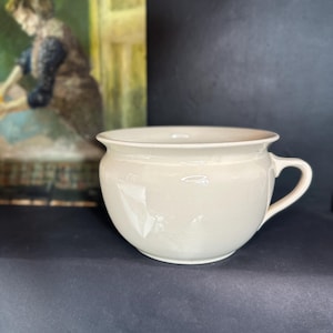 May include: A cream-coloured ceramic chamber pot with a wide, rounded bowl, a flared rim, and a single side handle. The surface is smooth and glossy, reflecting light, and the vessel features a deep, rounded base for stability.