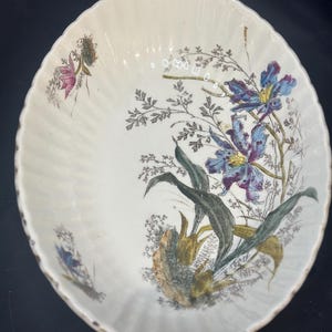 May include: An oval ceramic bowl with a cream-coloured base and a fluted, scalloped rim. The interior features a botanical illustration of blue and purple iris flowers, green leaves, and delicate sprigs of wildflowers in shades of brown and green.