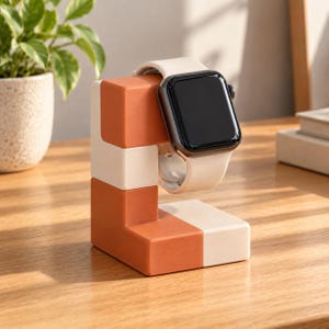 May include: A smartwatch with a black screen and a light beige silicone band is mounted on a block-shaped charging stand. The stand is constructed from interlocking matte terracotta orange and cream-colored rectangular segments.