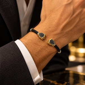 May include: A black rubber cuff bracelet featuring two gold-toned shield-shaped terminals. Each terminal is accented with small, embedded black stones arranged in a grid pattern, creating a textured and metallic contrast against the smooth band.