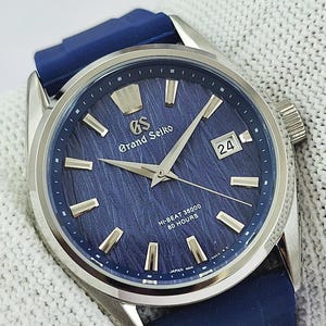 May include: A Grand Seiko watch with a silver stainless steel case and a dark blue silicone strap. The dial features a textured blue finish, silver-tone hour markers, silver hands, a date window displaying 24, and text reading GS Grand Seiko.