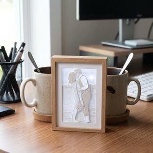 May include: A rectangular light wood frame containing a layered paper art piece depicting a couple kissing on a beach. The artwork features white and beige paper cutouts showing two figures embracing, with a light background and a simple wooden border.