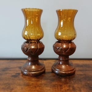 Peut inclure: Une paire de bougeoirs dotés d'abat-jours en verre ambré avec des nervures verticales. Chaque abat-jour repose sur une base en bois brun foncé présentant un motif géométrique en losange sculpté autour du centre du pied.