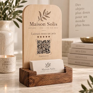 Peut inclure: Un panneau en bois clair à sommet arrondi, orné d'un motif de branche d'olivier, du texte Maison Solis, cinq étoiles et un QR code. Il repose sur un socle en bois foncé contenant des cartes de visite avec le même logo et texte.
