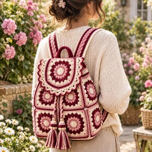 Puede incluir: Una mochila de ganchillo con un patrón de motivos cuadrados en tonos crema, rosa claro y burdeos intenso. La bolsa tiene una solapa circular, un asa superior, correas trenzadas y un cierre de cordón rematado con dos grandes borlas de hilo.