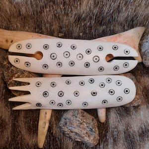 Könnte beinhalten: Zwei cremefarbene, längliche Haaraccessoires aus Knochenmaterial. Das obere Teil hat einen mittigen Schlitz und abgerundete Enden, das untere Teil ist mit vier Zinken versehen. Beide Objekte sind mit einem Muster aus kleinen, dunklen Kreisen verziert.
