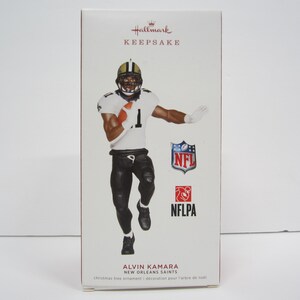 May include: A Hallmark Keepsake ornament featuring Alvin Kamara, a football player for the New Orleans Saints. The ornament is a detailed figurine of Kamara in his uniform, holding a football and running. The ornament is packaged in a box with the Hallmark logo and the NFLPA logo.