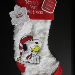 May include: White and red Christmas stocking with a white fluffy texture. The stocking has a red cuff with the text "Baby's First Christmas" embroidered on it. There is a white snowflake with the year "2019" embroidered on it. There is a white embroidered image of Snoopy holding Woodstock. The stocking has a red Hallmark tag with the text "Commemorative Stocking Peanuts" on it.