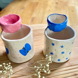 Könnte beinhalten: Zwei handgefertigte Keramikbecher mit unregelmäßigen Rändern. Der linke Becher ist cremefarben mit rosa Punkten und einem blauen Schmetterling. Der rechte Becher ist cremefarben mit kleinen blauen Blumen. Jeder Becher hat einen kleinen, schalenartigen Rand.