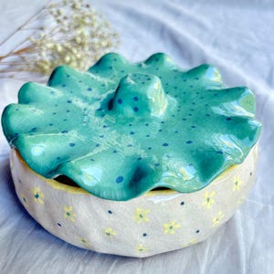 Op de afbeelding: Een ronde keramische pot met deksel en een craquelé glazuurafwerking. De basis is gebroken wit met kleine gele bloemen en donkerblauwe stippen. Het deksel is een gegolfde, turquoise schijf met een centrale knop en blauwe stippen.