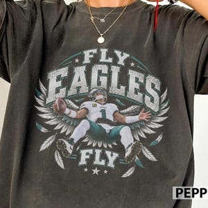 May include: A charcoal grey t-shirt featuring a graphic of a football player in a white jersey with the number 1, holding a football. The design includes large white text reading FLY EAGLES FLY, surrounded by stylised wings and feathers in white.