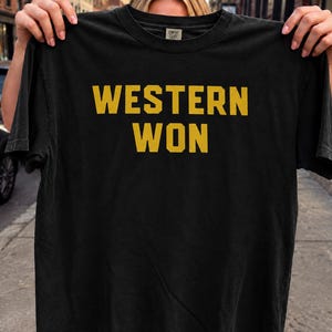 Puede incluir: Una camiseta negra de manga corta y cuello redondo con el texto WESTERN WON impreso en letras mayúsculas amarillas en negrita en el pecho. La prenda tiene un corte estándar con un cuello acanalado y una pequeña etiqueta de marca en el interior.