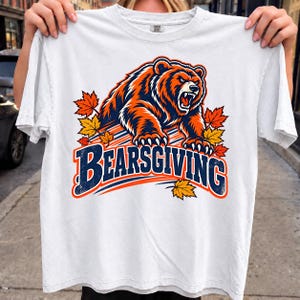 May include: A white short-sleeve t-shirt featuring a graphic of a snarling brown bear with orange and navy blue accents. The design includes scattered autumn maple leaves and the text "BEARSGIVING" in bold navy blue letters with an orange outline.