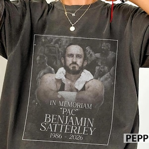 Puede incluir: Una camiseta gris carbón con un estampado gráfico en blanco y negro de un hombre barbudo con una toalla blanca alrededor del cuello. El texto dice: IN MEMORIAM "PAC" BENJAMIN SATTERLEY 1986 - 2026. El diseño está enmarcado por un borde blanco.