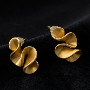 May include: A pair of gold-toned stud earrings featuring a sculptural, wavy, ribbon-like design with a matte finish. Each earring has a post back with a clear plastic clutch, showcasing a fluid, curved shape that creates depth and dimension.