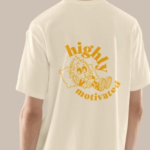 Könnte beinhalten: Ein cremefarbenes Kurzarm-T-Shirt mit einer gelben Grafik auf der Rückseite. Die Grafik zeigt eine Zeichentrickfigur, die auf einem Kissen liegt, wobei der Text "highly motivated" in einer runden, fetten Schrift um die Illustration gebogen ist.