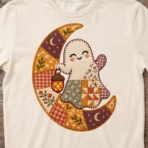 Può includere: Una t-shirt color crema con la grafica di un fantasma sorridente che tiene in mano una lanterna a forma di ghianda mentre siede su una luna crescente. La luna e il fantasma presentano un motivo patchwork con fiori, quadretti e stelle.